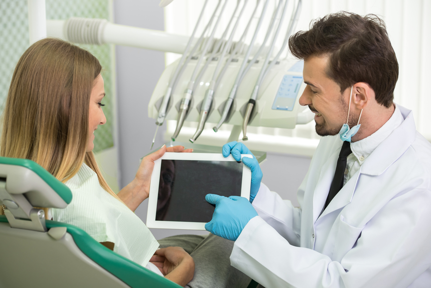 fotografia-de-dentista-sorridente-mostrando-informações-no-tablet-para-paciente-em-consultório-odontológico